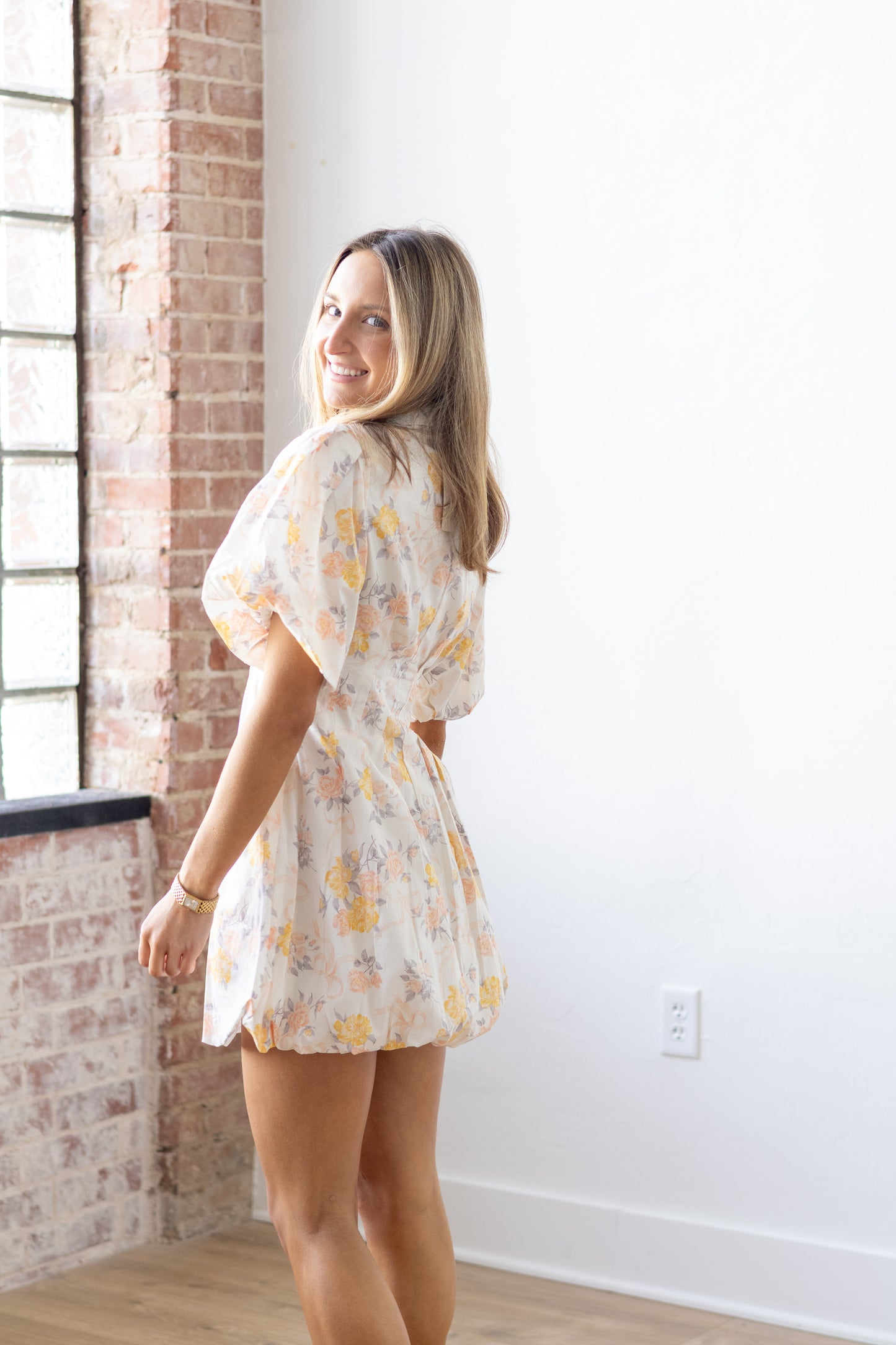 Woman wearing a floral dress standing in a room with brick walls and a white wall.