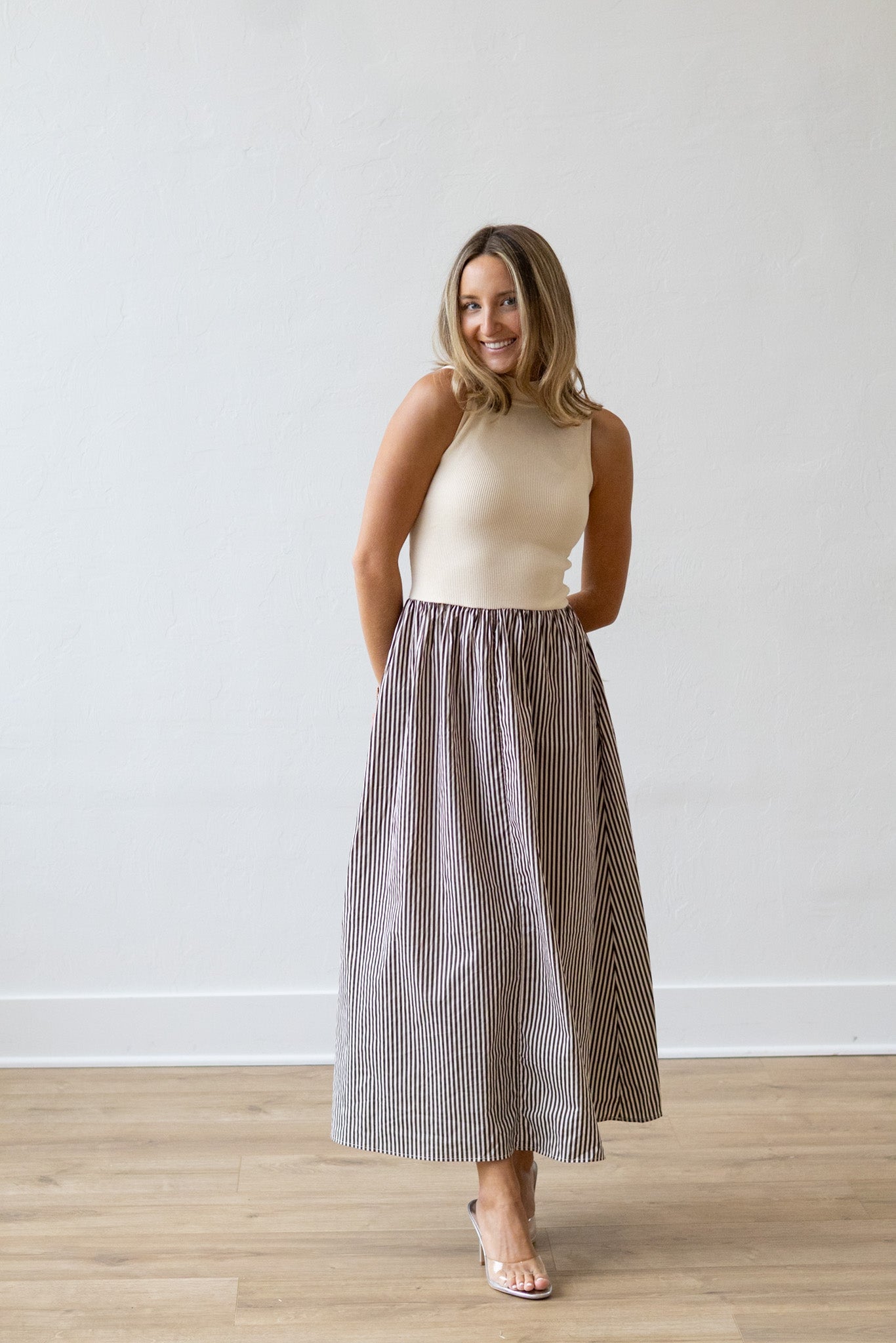 Woman wearing a beige top and striped skirt standing against a white wall.