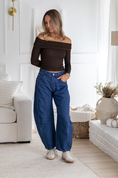Woman in a living room wearing a brown off-shoulder top and blue jeans.