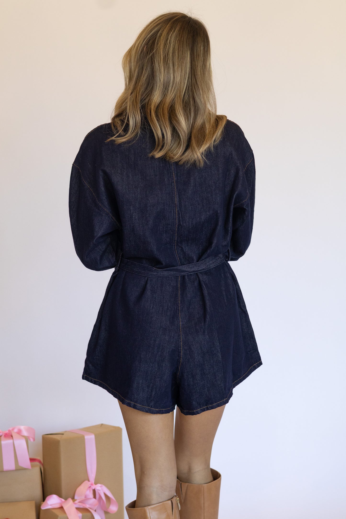 Person wearing a navy denim romper with brown boots and gift boxes in the foreground.