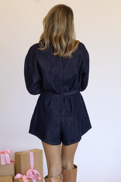 Person wearing a navy denim romper with brown boots and gift boxes in the foreground.