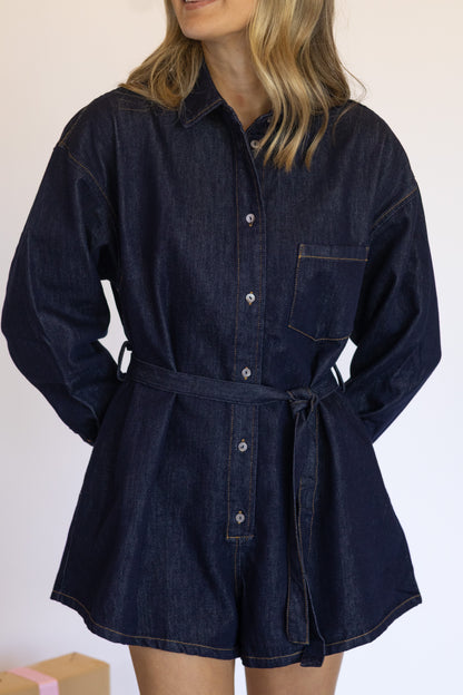 Woman wearing a dark denim romper with a belt against a plain background