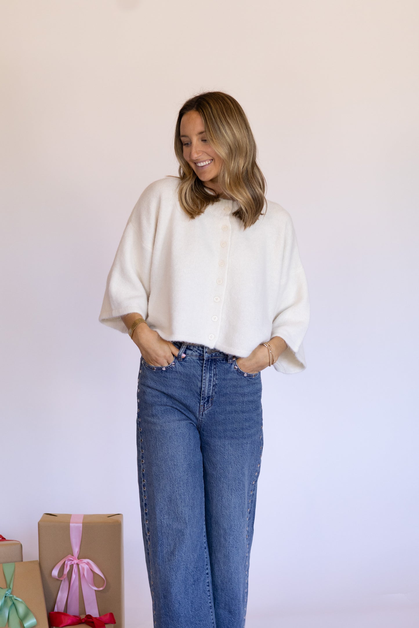 Woman wearing a white blouse and blue jeans standing against a plain background with gift boxes.