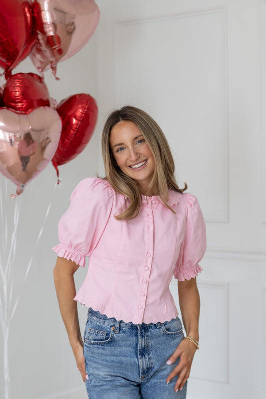 Woman in a pink blouse and jeans standing next to heart-shaped balloons against a white wall.