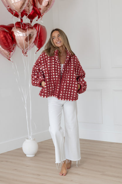 Woman in a red checkered shirt and white pants standing next to heart-shaped balloons indoors.