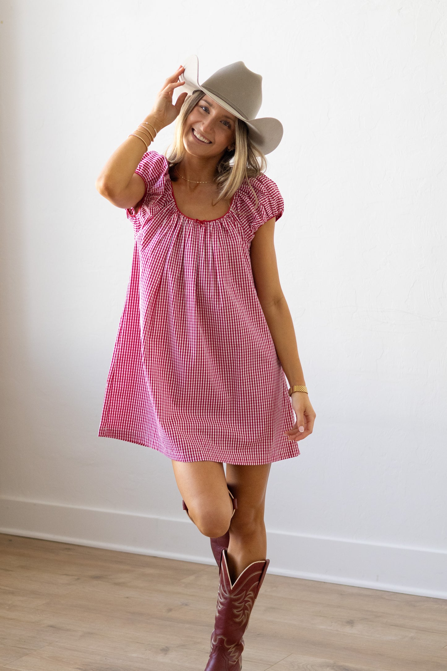 Woman wearing a pink checkered dress, beige hat, and red cowboy boots against a white wall.
