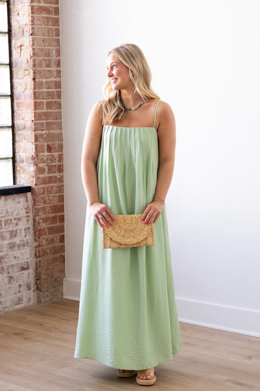 Woman in a light green dress holding a straw clutch against a white wall with brick accents.