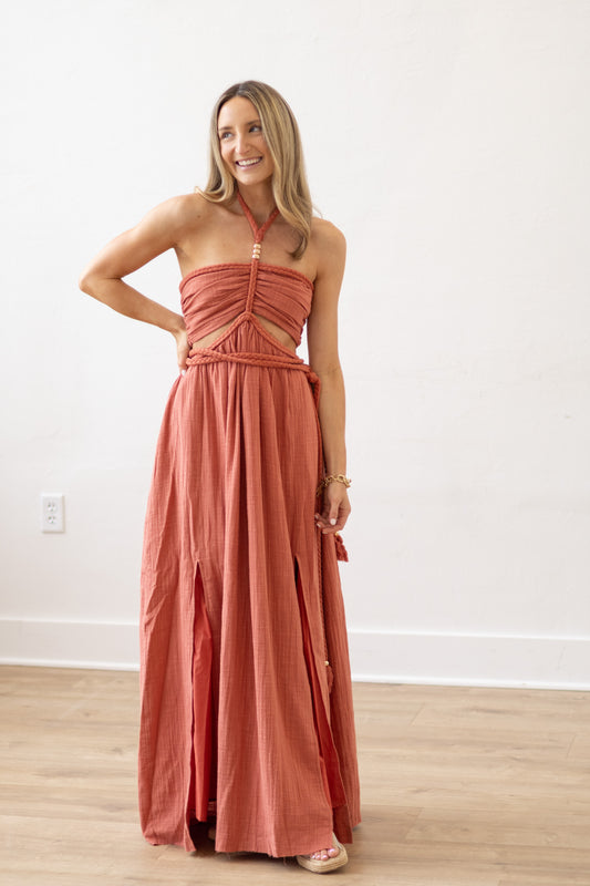 Woman wearing a long coral dress standing in a room with a white wall and wooden floor.