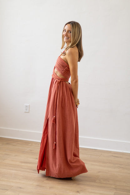Woman wearing a long rust-colored dress standing in a room with wooden flooring and a white wall.