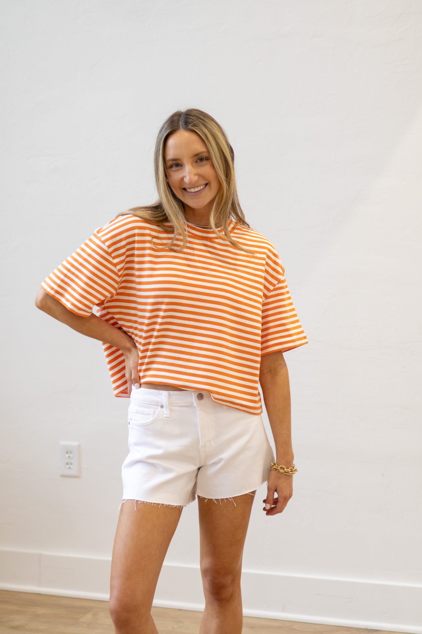 Woman wearing an orange and white striped shirt and white shorts standing against a plain white wall.