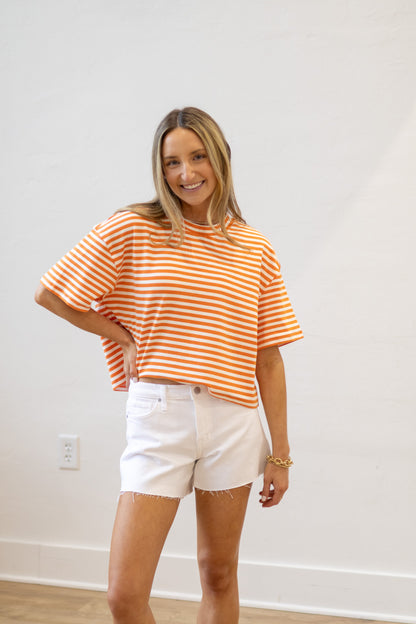 Woman wearing an orange and white striped shirt and white shorts standing against a plain white wall.
