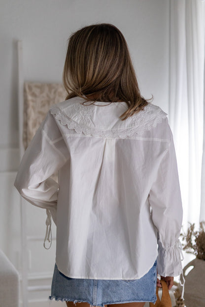 Woman wearing a white lace-trimmed blouse in a softly lit room.