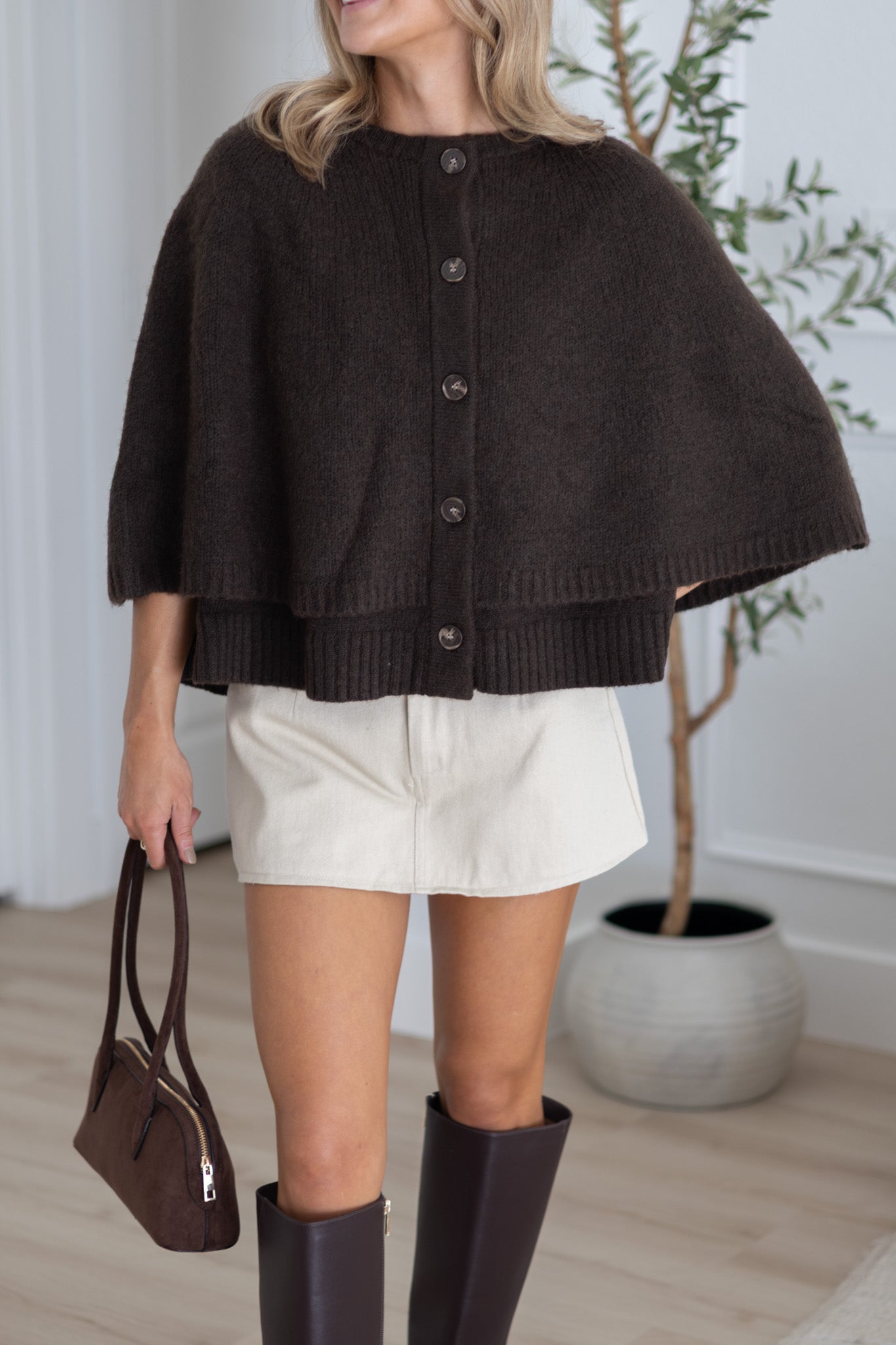 Woman wearing a brown knitted cape over a black top, white shorts, and brown boots, holding a brown handbag.