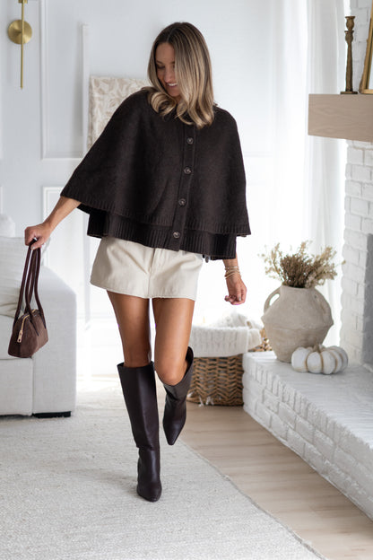Woman in a black cape and white shorts walking in a living room.