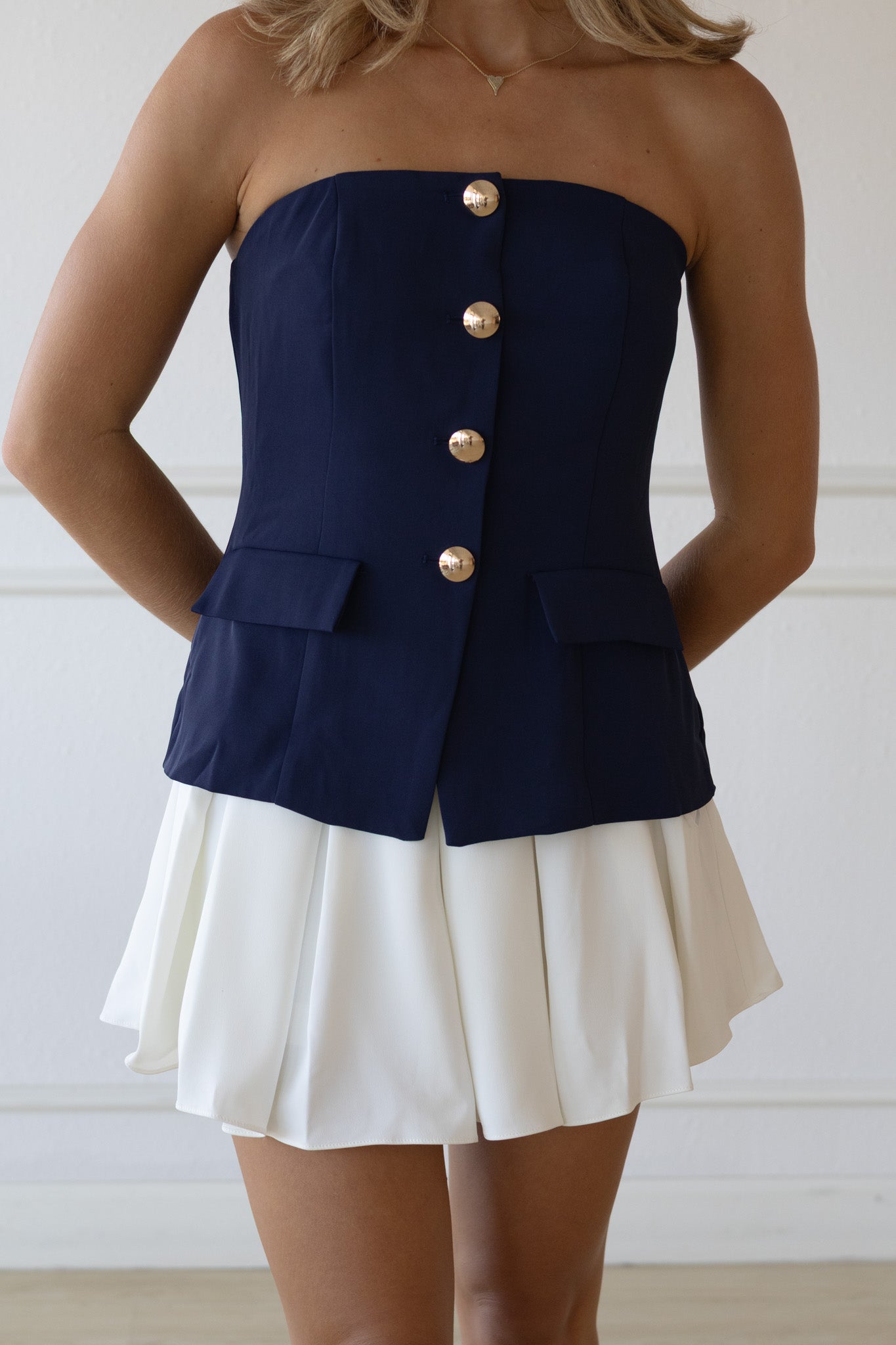 Woman wearing a navy strapless top with gold buttons and a white skirt against a white background