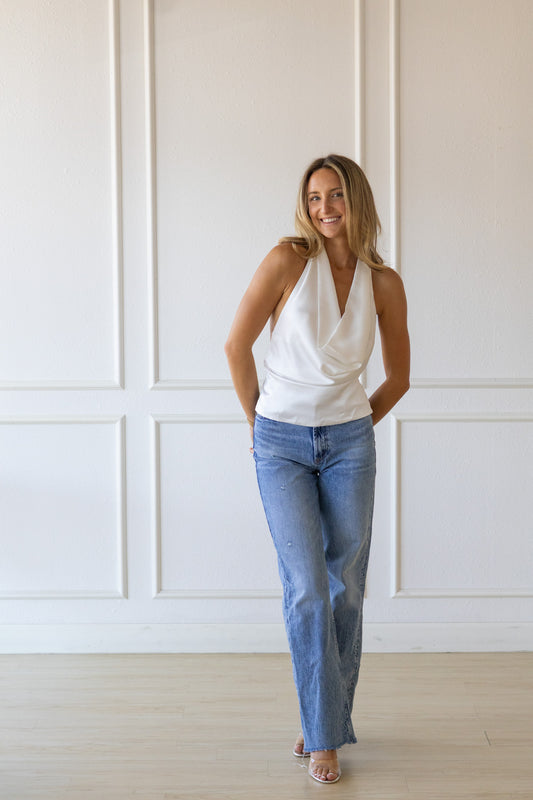 Woman wearing a white sleeveless top and blue jeans standing against a white paneled wall.