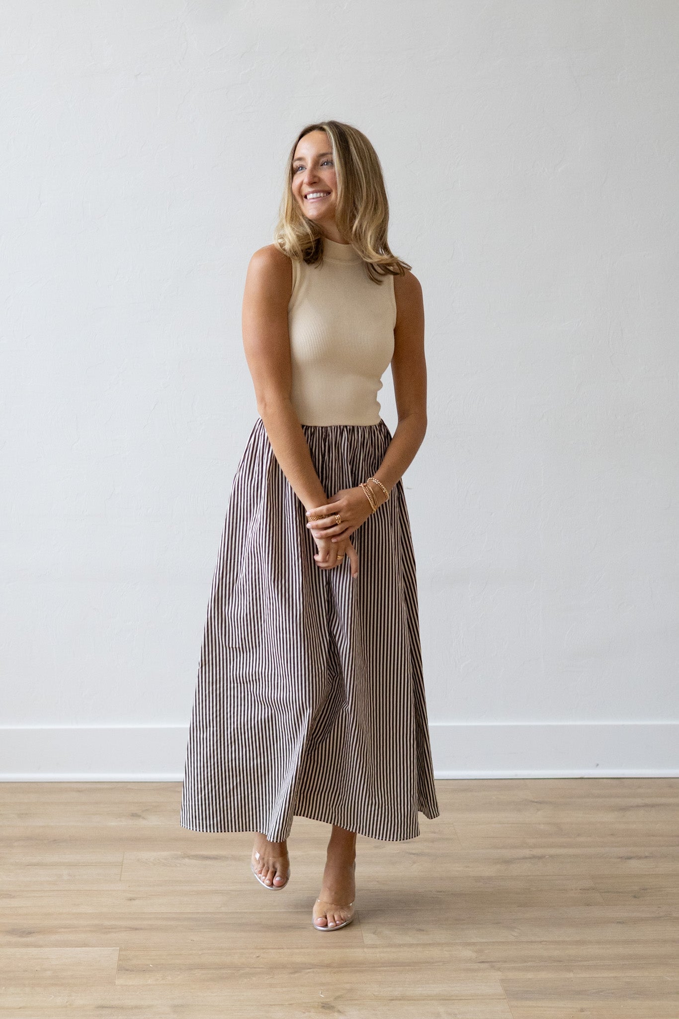 Woman wearing a beige top and striped skirt standing against a white wall.
