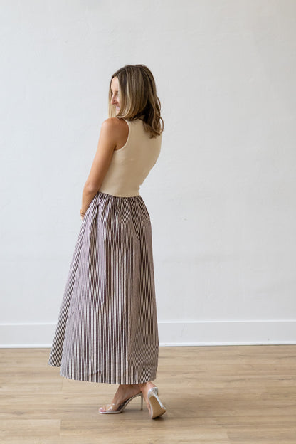 Woman wearing a beige dress with a sleeveless top and striped skirt against a white wall.