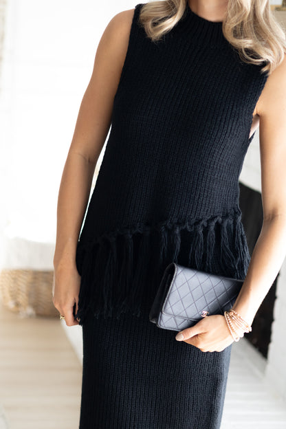 Woman wearing a black sleeveless knit top with ruffled hem, holding a gray clutch.