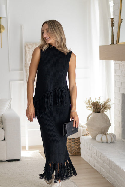 Woman wearing a black sleeveless dress with fringe details in a living room setting.