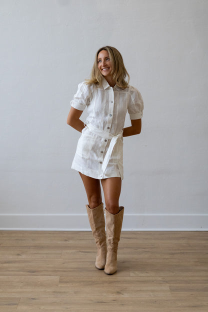 Woman wearing a white dress with buttons and beige knee-high boots standing against a plain wall.