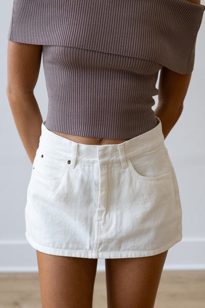 Person wearing a brown ribbed top and white denim skirt against a plain background
