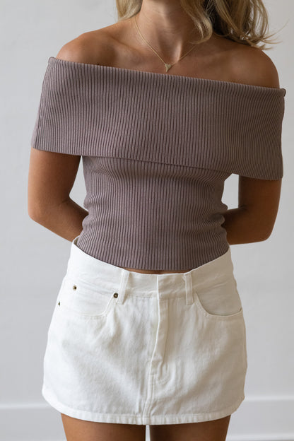 Person wearing an off-shoulder brown top and white denim skirt against a plain background