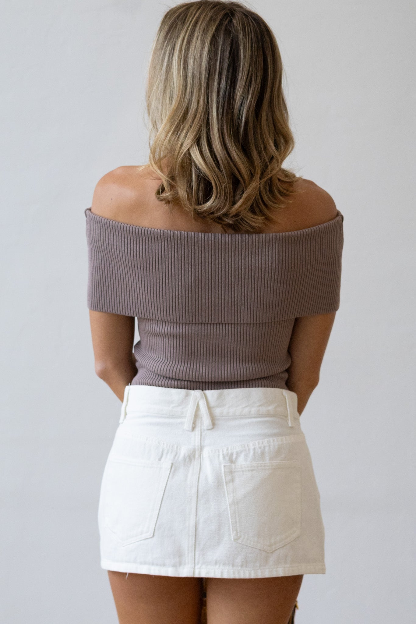Woman wearing a brown off-shoulder top and white skirt against a plain background