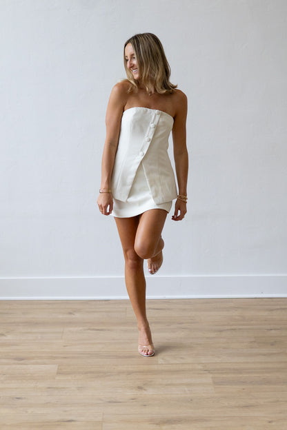 Woman wearing a white strapless dress standing on a wooden floor with a plain white wall background