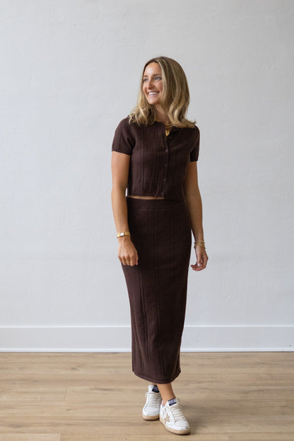 Woman wearing a brown outfit standing against a white wall.