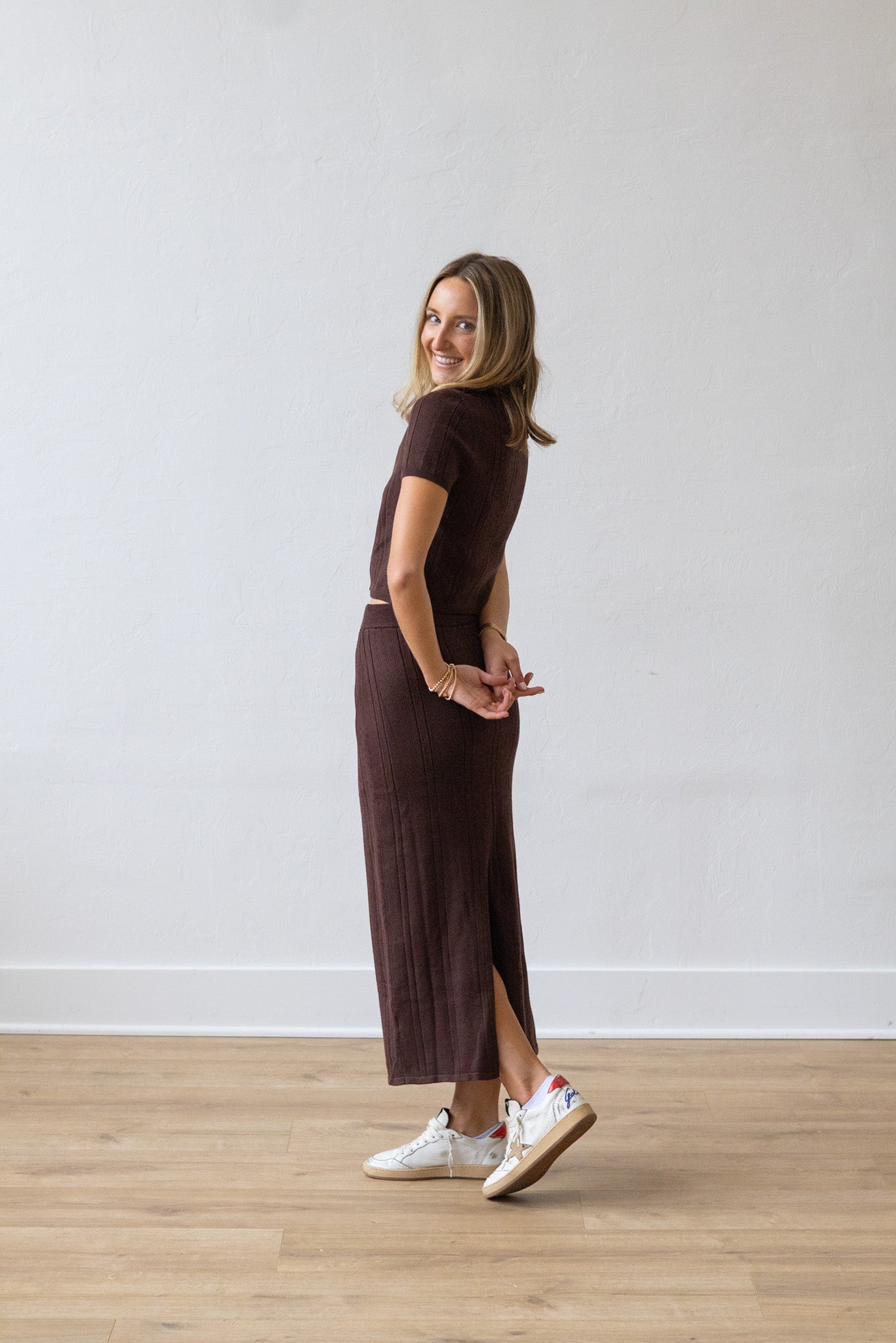 Woman wearing a brown dress standing on a wooden floor with a white wall background
