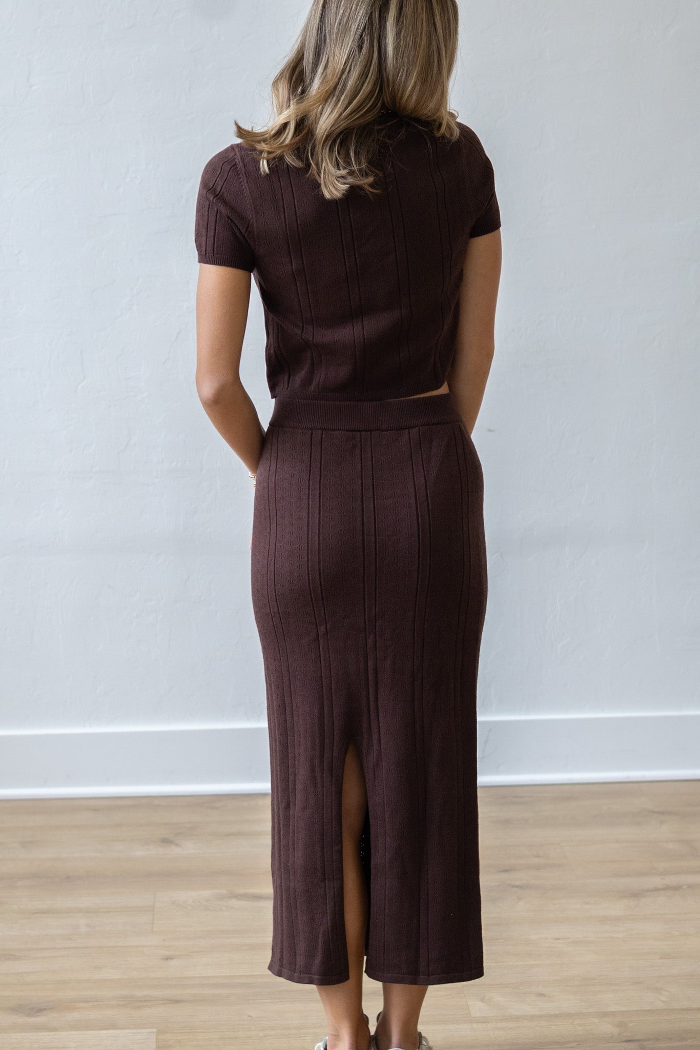 Person wearing a brown outfit with a skirt and top, standing against a white wall.