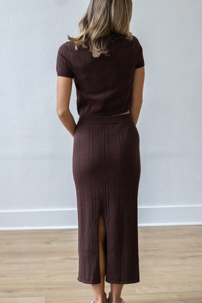 Person wearing a brown outfit with a skirt and top, standing against a white wall.