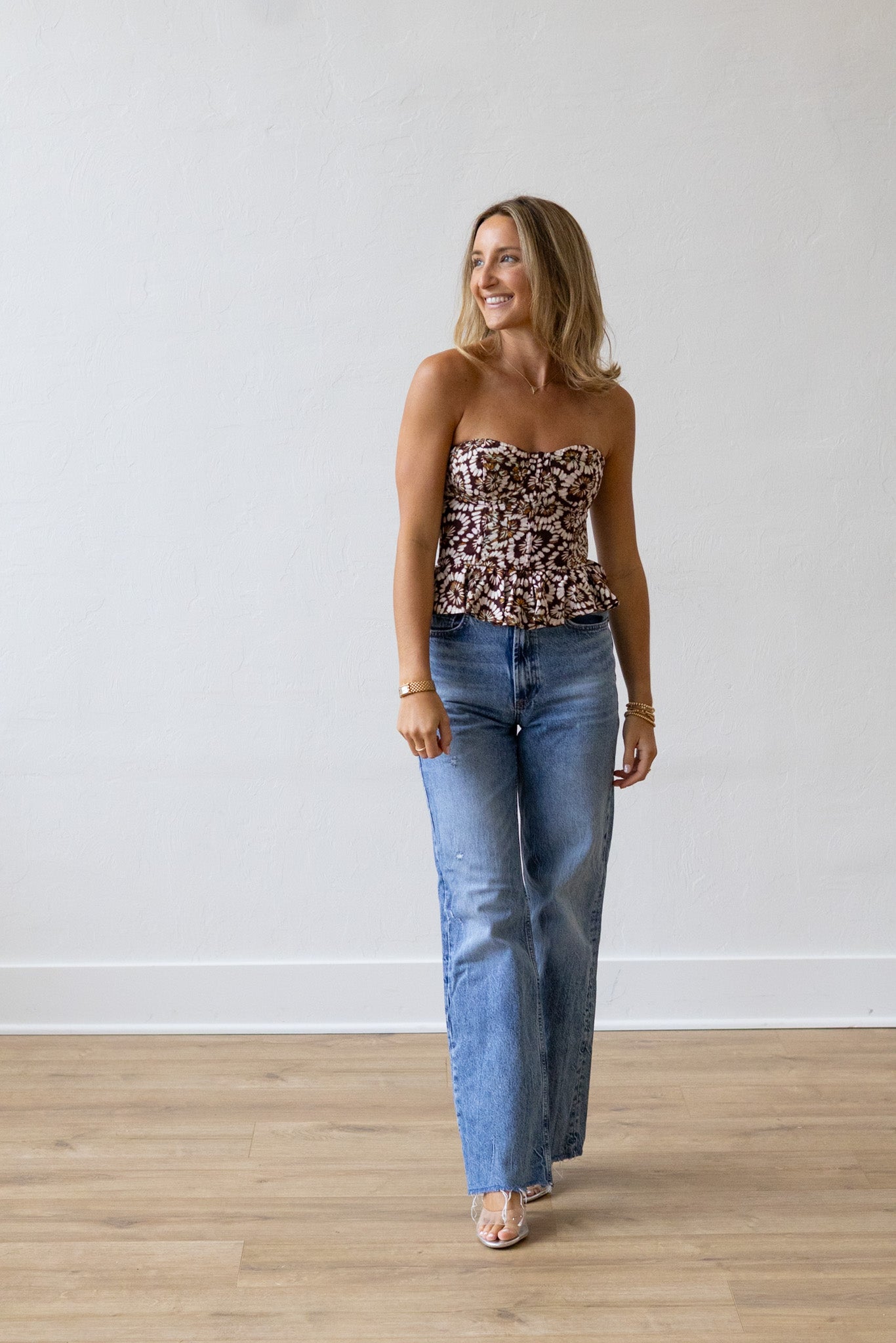 Woman wearing a patterned top and blue jeans standing against a white wall.