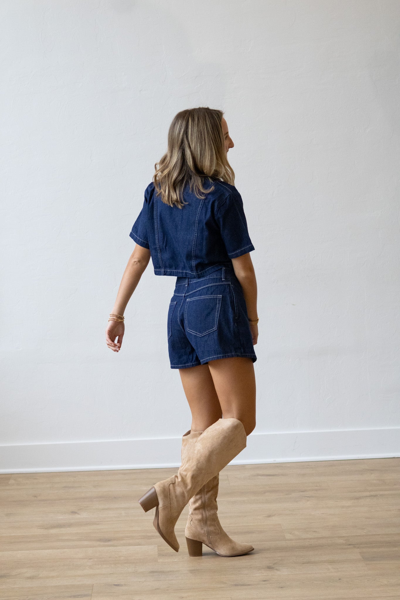 Woman wearing a denim outfit with beige boots against a white wall.