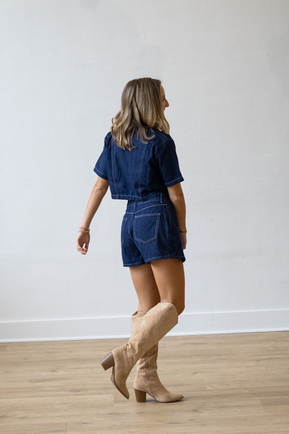 Woman wearing a denim outfit with beige boots against a white wall.