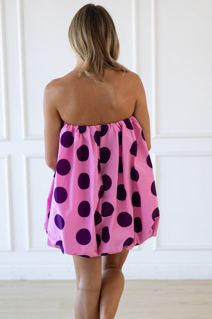 Woman wearing a pink strapless dress with purple polka dots against a white wall.