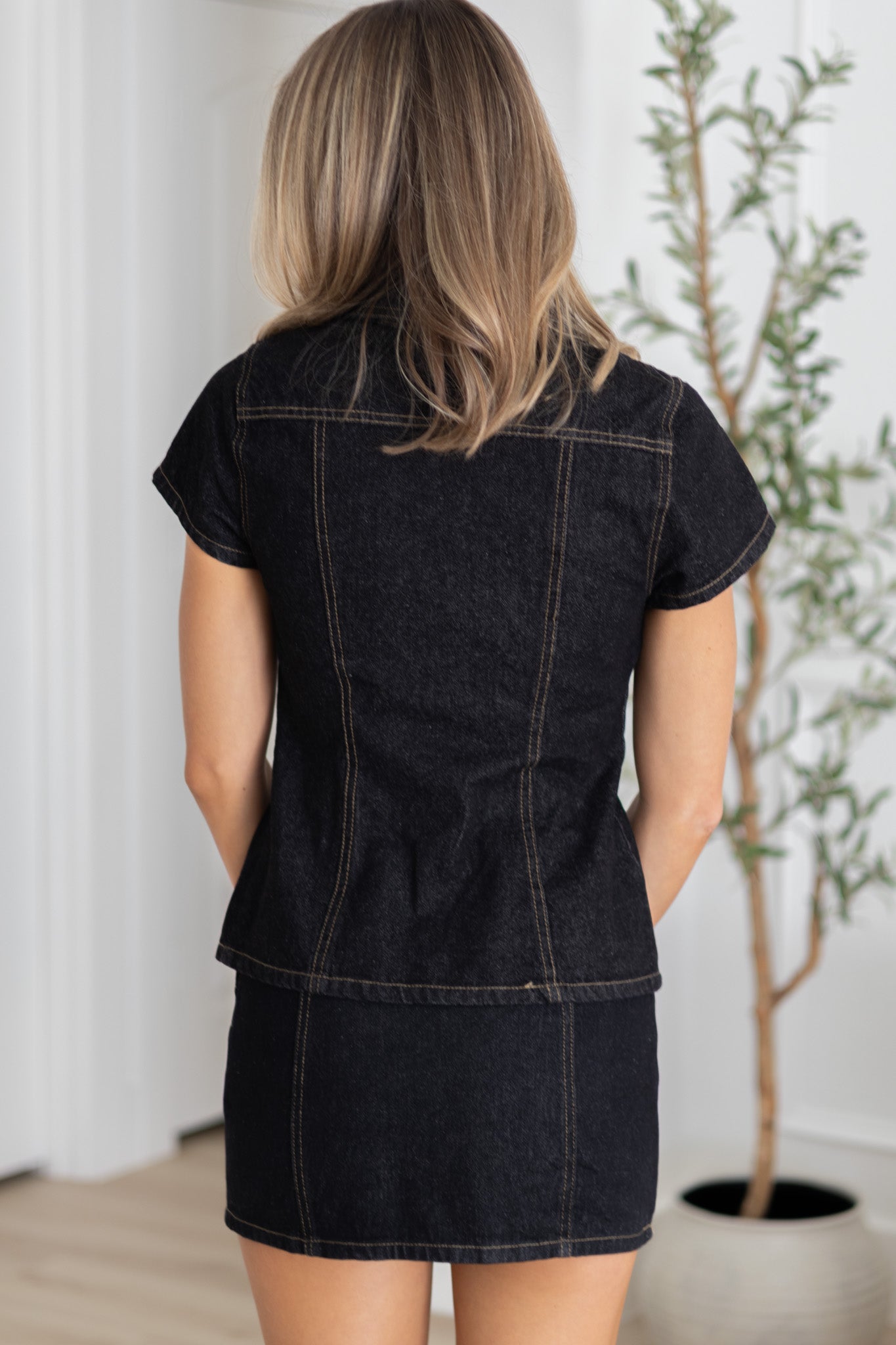 Woman wearing a black denim dress standing in a room with a plant.