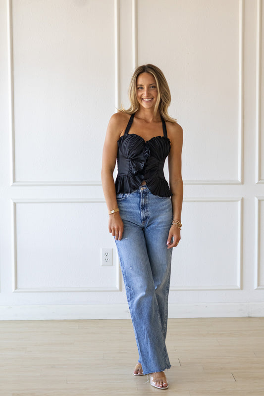 Woman wearing a black top with floral details and blue jeans against a white paneled wall.